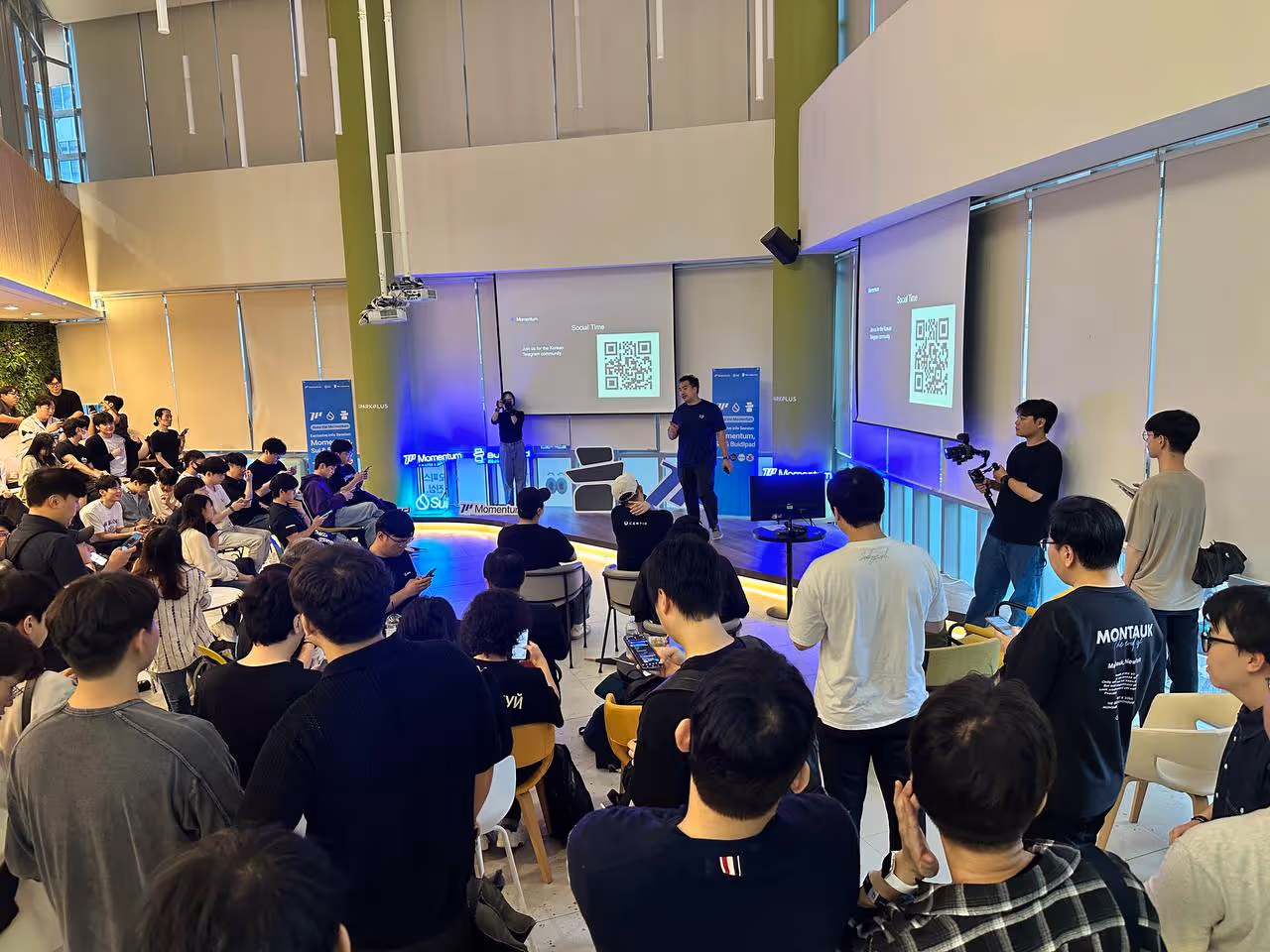 Crowded tech event with speaker on stage, audience using phones, and a large QR code on screen.