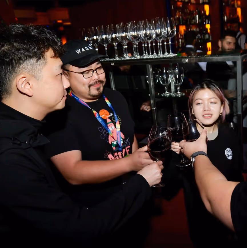Three people holding wine glasses in a toast at a bar with glasses on a shelf behind them.