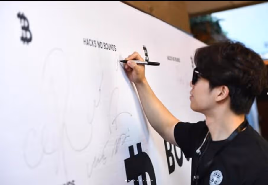 Person wearing sunglasses signing a large white board with Bitcoin symbols and 'PLACE NO BOUNDS' text.