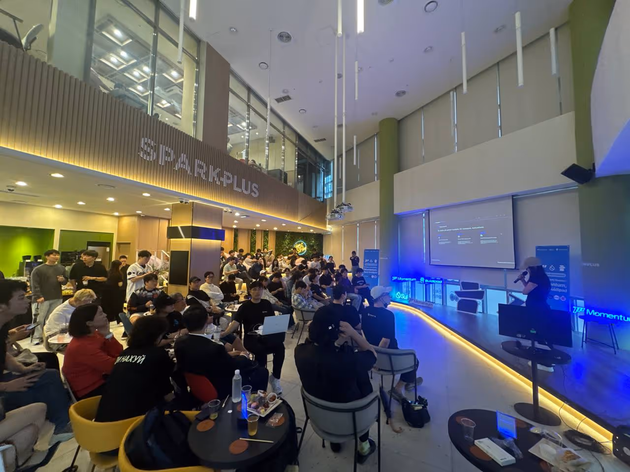 Crowded modern event space with seated audience listening to presenter near a large screen.