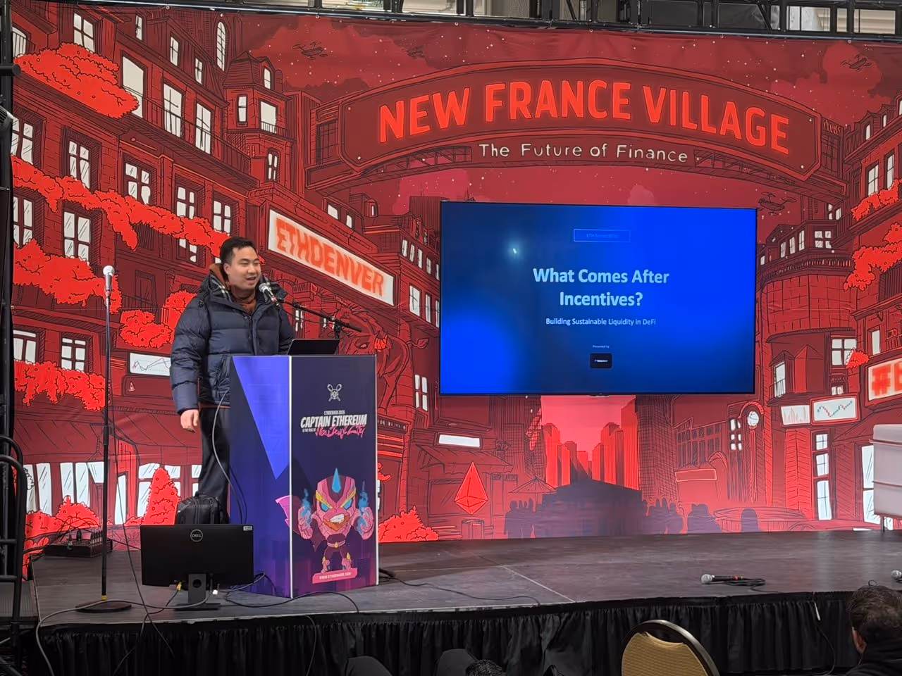 Speaker in winter coat at podium presenting on building sustainable liquidity in DeFi at ETH Denver.