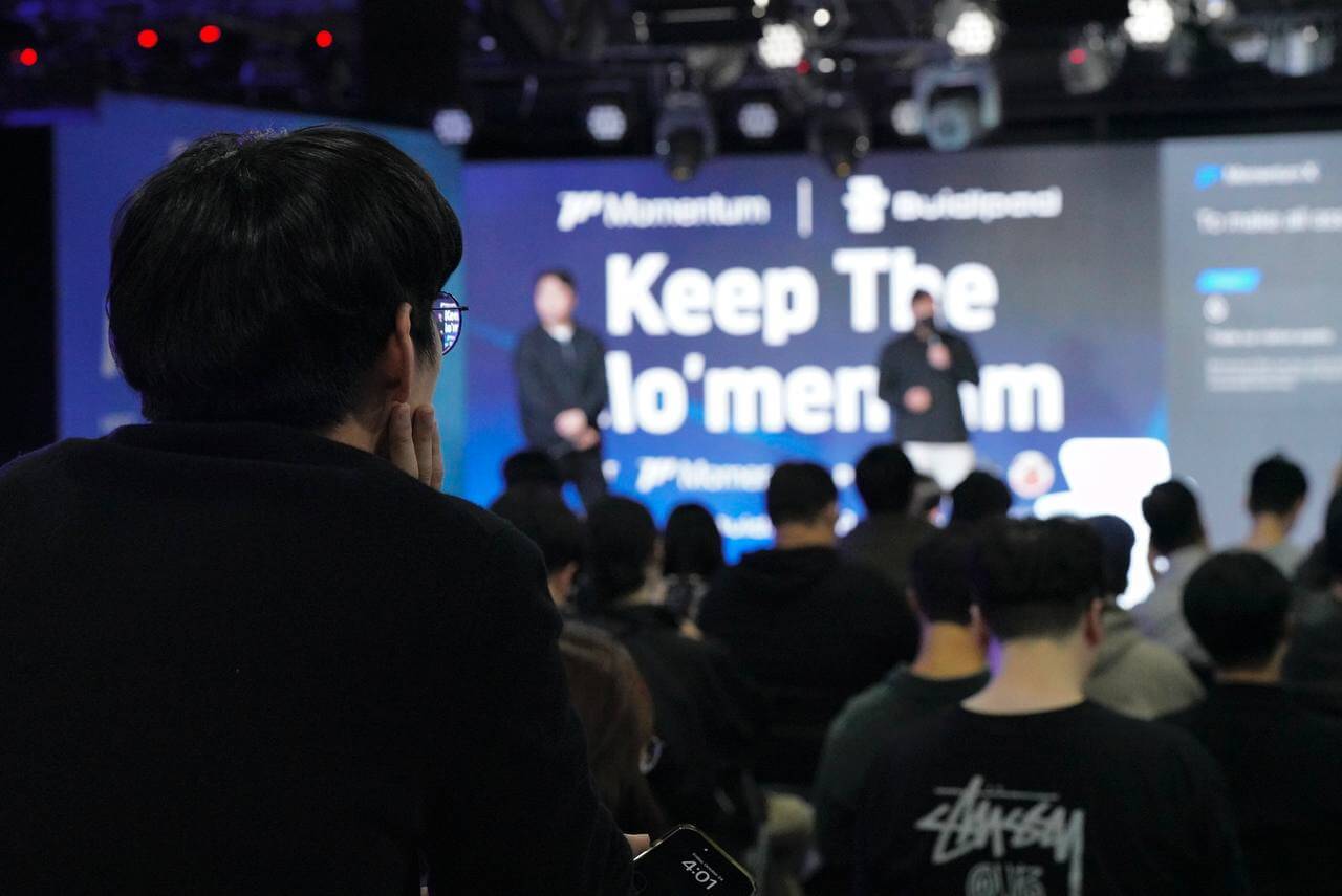 Audience watching two presenters on stage with a screen showing the phrase 'Keep The Momentum'.